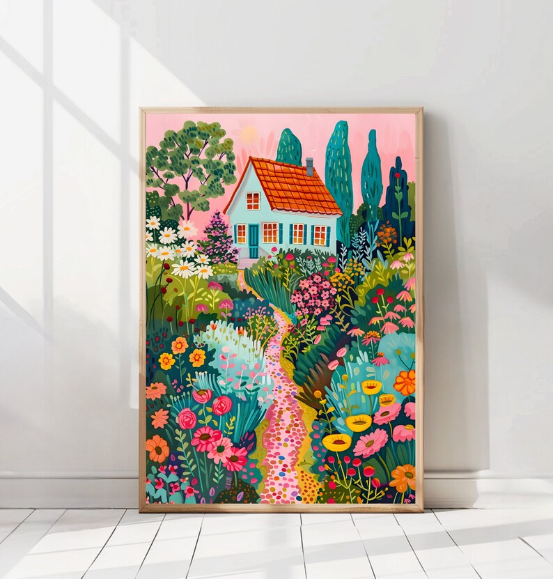May include: A framed art print showing a garden scene. A small house with a red tiled roof and blue shutters is surrounded by colourful flowers and greenery. A stone path winds through the garden, set against a pink sky. The artwork is in a wooden frame.
