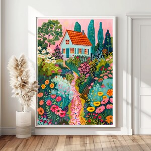 May include: A framed art print showing a garden path leading to a small house with a red tiled roof. The artwork features vibrant flowers, trees, and a pink sky, creating a cheerful scene. The print is displayed in a white frame, suitable for home decor.