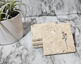 May include: A stack of square, beige stone coasters with a delicate pink floral design. A potted air plant with long, green leaves sits to the left. The background is a marble-patterned surface.