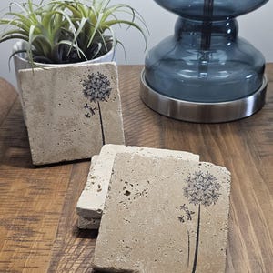 May include: Set of four beige stone coasters with a black dandelion design. Each coaster features a dandelion with seeds blowing in the wind.