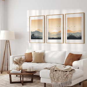 May include: Three framed landscape prints with a sunset over mountains and birds flying in the sky. The prints are in a living room setting with a white couch and a wooden coffee table.