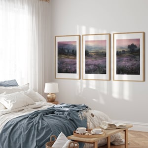 May include: Three framed landscape paintings depicting a purple field with a distant view of a town and hills. The paintings are hung on a white wall in a bedroom setting.