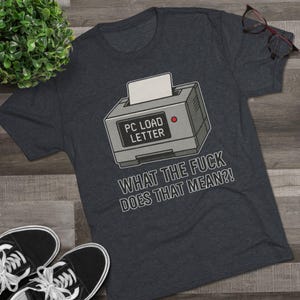May include: Dark gray t-shirt featuring a retro computer printer graphic with the text "PC LOAD LETTER" and the question "WHAT THE FUCK DOES THAT MEAN?!" in white. The shirt has a crew neck and short sleeves.