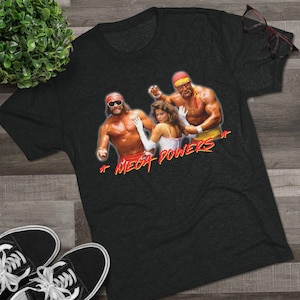 May include: Dark gray t-shirt featuring a graphic of three people with the text "MEGA POWERS" in red and yellow. The graphic includes a woman in a white dress and two men. The shirt is on a wooden surface.
