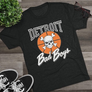 Könnte beinhalten: Dunkelgraues T-Shirt mit dem Schriftzug "DETROIT" über einem Basketball mit einem Totenkopf und gekreuzten Knochen. Die Worte "Bad Boys" stehen unter dem Basketball. Schwarze und weiße Turnschuhe befinden sich in der unteren linken Ecke.