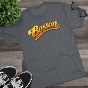 May include: A heather gray t-shirt with the word "Boston" in a retro, yellow and orange script. The shirt is displayed on a wooden surface, with black sneakers and eyeglasses visible.