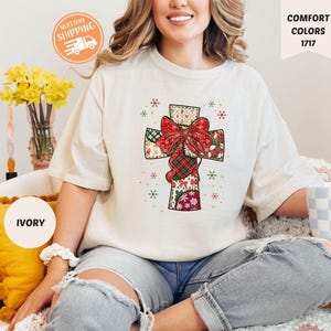 Puede incluir: Camiseta de color marfil con un diseño de cruz colorida con un lazo rojo. La cruz está decorada con patrones de patchwork y copos de nieve. La camiseta forma parte de la colección Comfort Colors.