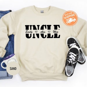 May include: A beige sweatshirt with the word "UNCLE" printed in black bold letters. The names "Emily", "Alex", and "Mia" are printed below the word "UNCLE" with a heart symbol between each name.