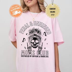 Puede incluir: Camiseta rosa claro con las palabras "FERAL & UNHINGED" sobre un dise&ntilde;o de calavera y el texto "MOMS CLUB" debajo. La camiseta tambi&eacute;n tiene el texto "FUELED BY CAFFIENE & CHAOS".