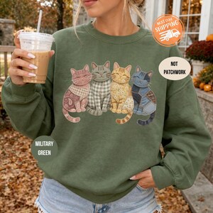 May include: A military green crewneck sweatshirt featuring a graphic of four cats with varied patterns including gingham, floral, and patchwork designs. The text "NOT PATCHWORK" is printed on the right side of the chest area above the cats.