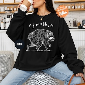 May include: A black long-sleeve sweatshirt featuring a white graphic illustration of a raccoon in profile. Above the raccoon, the name jimothy is printed in white lowercase letters, flanked by two small white heart outlines on each side.