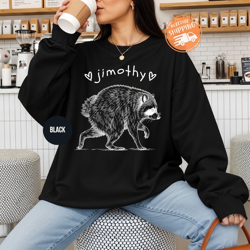 May include: A black long-sleeve sweatshirt featuring a white graphic illustration of a raccoon in profile. Above the raccoon, the name jimothy is printed in white lowercase letters, flanked by two small white heart outlines on each side.