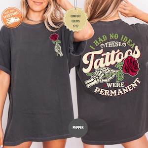 May include: A charcoal gray short-sleeve t-shirt featuring a graphic of a skeleton hand holding a red rose on the front left chest. The back displays a larger version of the same graphic with the text I HAD NO IDEA THESE TATTOOS WERE PERMANENT.