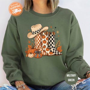 May include: A military green crewneck sweatshirt featuring a graphic print of a tan cowboy hat, two patterned cowboy boots, and several orange pumpkins. The boots display checkered and pumpkin-themed designs, accented by small star and leaf illustrations.