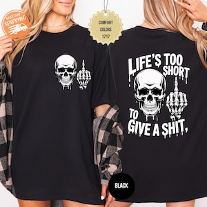 May include: Black t-shirt featuring a white skull graphic with a middle finger gesture. The back of the shirt displays a larger skull and the text "LIFE'S TOO SHORT TO GIVE A SHIT." The shirt is a Comfort Colors 1717.