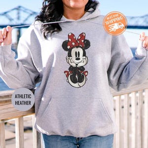 May include: Athletic heather gray hoodie featuring a Minnie Mouse graphic with a red bow and polka dots. The hoodie has a front pocket and a drawstring hood. The text "ATHLETIC HEATHER" is printed on the bottom left.