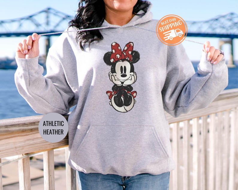 May include: Athletic heather gray hoodie featuring a Minnie Mouse graphic with a red bow and polka dots. The hoodie has a front pocket and a drawstring hood. The text "ATHLETIC HEATHER" is printed on the bottom left.