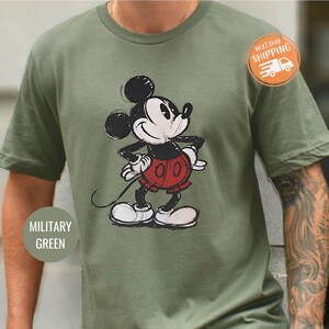 May include: Military green t-shirt featuring a sketched Mickey Mouse graphic in black, red, and white. The shirt has a classic crew neck and short sleeves. The text "MILITARY GREEN" is on the bottom left of the shirt.
