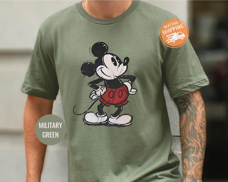 May include: Military green t-shirt featuring a sketched Mickey Mouse graphic in black, red, and white. The shirt has a classic crew neck and short sleeves. The text "MILITARY GREEN" is on the bottom left of the shirt.
