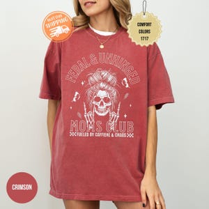 Puede incluir: Camiseta de color carmes&iacute; con el texto "FERAL & UNHINGED MOMS CLUB" y un dise&ntilde;o de calavera. La camiseta tambi&eacute;n tiene la frase "FUELED BY CAFFIENE & CHAOS". La camiseta tiene mangas cortas y cuello redondo.