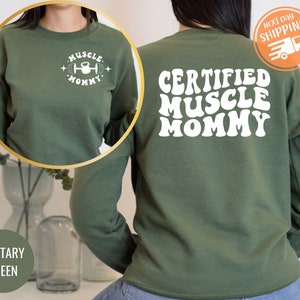 Op de afbeelding: Groen sweatshirt met de tekst "Certified Muscle Mommy" in witte retro-lettertype op de achterkant en "Muscle Mommy" met een halter- en kettlebell-afbeelding op de voorkant.