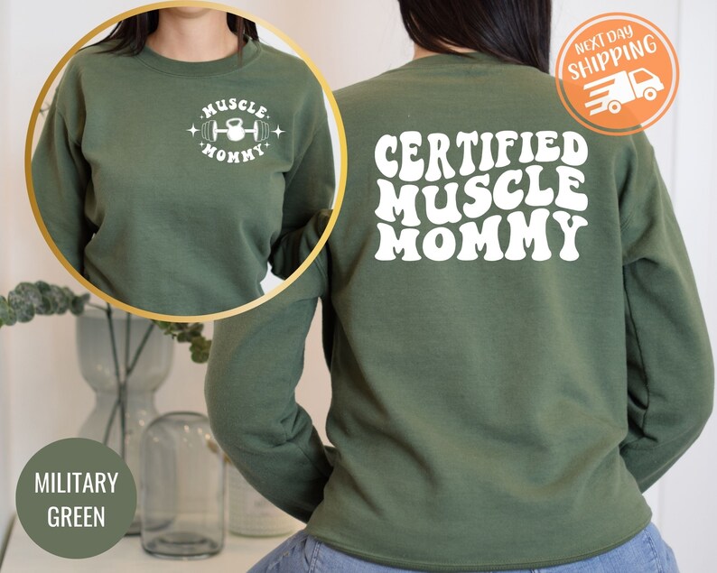 Op de afbeelding: Groen sweatshirt met de tekst "Certified Muscle Mommy" in witte retro-lettertype op de achterkant en "Muscle Mommy" met een halter- en kettlebell-afbeelding op de voorkant.