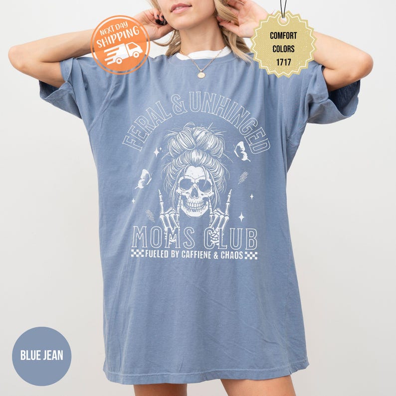 Puede incluir: Camiseta azul vaquero con texto y gr&aacute;ficos blancos. La camiseta presenta las palabras "FERAL & UNHINGED" sobre una calavera con un mo&ntilde;o desordenado y las palabras "MOMS CLUB" debajo. La camiseta tambi&eacute;n tiene el texto "FUELED BY CAFFIENE & CHAOS".