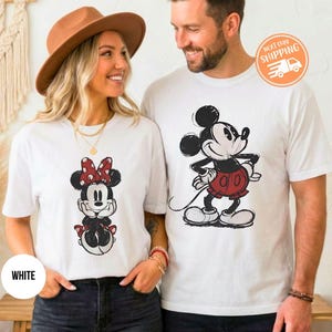 May include: White t-shirts featuring Mickey and Minnie Mouse designs. The Minnie Mouse shirt has a red polka dot bow. The Mickey Mouse shirt has a red skirt. The shirts are worn by a couple.
