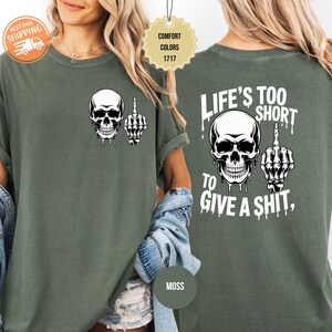 May include: Moss green t-shirt featuring a white skull graphic with a middle finger on the front. The back of the shirt displays a larger skull graphic and the text "LIFE'S TOO SHORT TO GIVE A SHIT,".