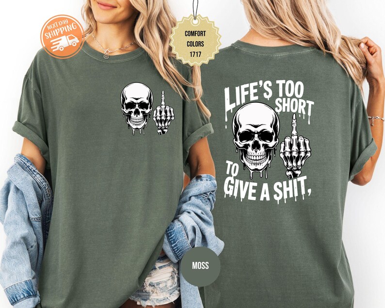 May include: Moss green t-shirt featuring a white skull graphic with a middle finger on the front. The back of the shirt displays a larger skull graphic and the text "LIFE'S TOO SHORT TO GIVE A SHIT,".