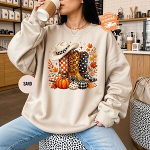 Könnte beinhalten: Ein sandfarbenes Sweatshirt mit Rundhalsausschnitt und einem Grafikdruck, der einen Cowboyhut, drei Westernstiefel mit Karomuster, Leopardenmuster und Punkten sowie drei Kürbisse zeigt, umgeben von Herbstblättern und kleinen Sternen.