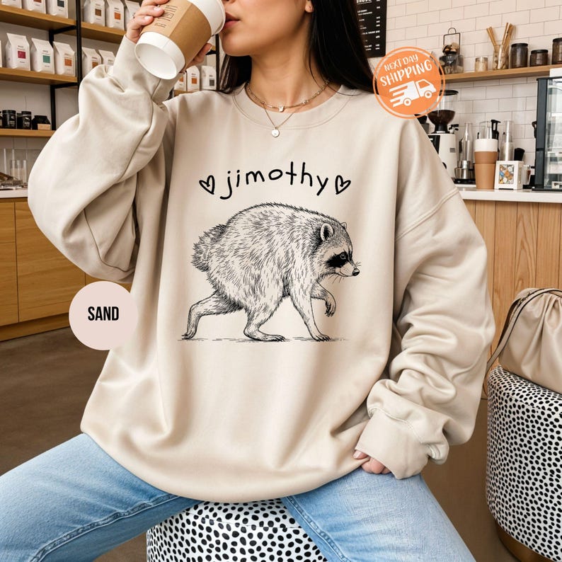 May include: A sand-colored crewneck sweatshirt featuring a black ink-style illustration of a raccoon walking. Above the raccoon, the text reads "jimothy" flanked by two small heart symbols. The garment has long sleeves and a ribbed neckline.