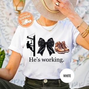Camiseta &quot;Ele está trabalhando&quot;, camiseta de eletricista, presente para esposa de eletricista, camiseta engraçada de eletricista, moletom com laço coquete, camiseta de esposa de colarinho azul