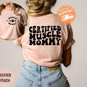 Op de afbeelding: Een heideperzikkleurig T-shirt met de tekst "CERTIFIED MUSCLE MOMMY" in zwarte retro-lettertypen. Het shirt wordt door een persoon gedragen.