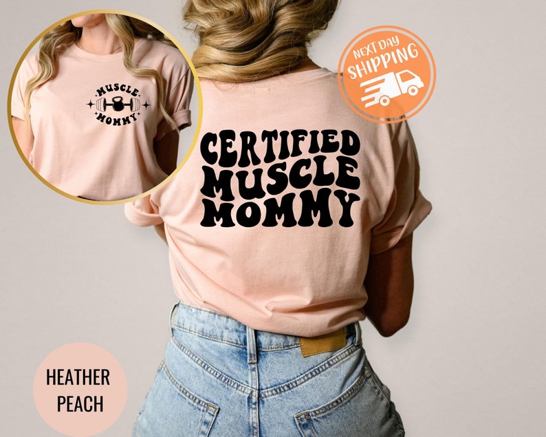 Op de afbeelding: Een heideperzikkleurig T-shirt met de tekst "CERTIFIED MUSCLE MOMMY" in zwarte retro-lettertypen. Het shirt wordt door een persoon gedragen.