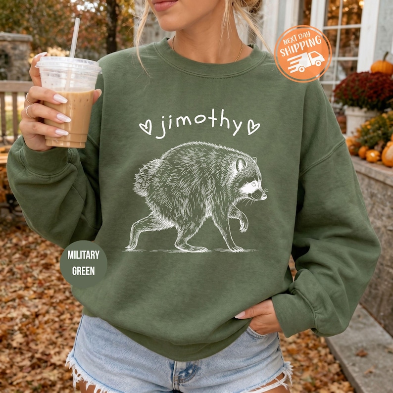 May include: A military green crewneck sweatshirt featuring a white line art illustration of a raccoon in the center. Above the raccoon, the text "jimothy" is printed in a lowercase white font, flanked by two small white heart outlines.