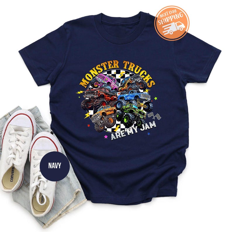 Puede incluir: Camiseta azul marino con un gr&aacute;fico colorido de monster trucks y el texto "MONSTER TRUCKS ARE MY JAM". El dise&ntilde;o incluye varios monster trucks de diferentes colores, sobre un fondo a cuadros. La camiseta se muestra con zapatillas blancas.