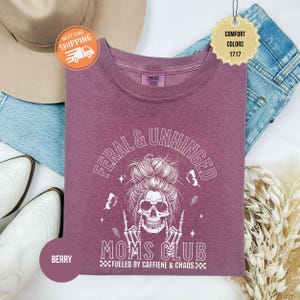 Puede incluir: Sudadera de color bayas con el texto "FERAL & UNHINGED MOMS CLUB" y un gr&aacute;fico de calavera. La camisa tambi&eacute;n tiene el texto "FUELED BY CAFFIENE & CHAOS". La camisa es de la marca Comfort Colors. La imagen incluye un par de jeans, un sombrero y botas.