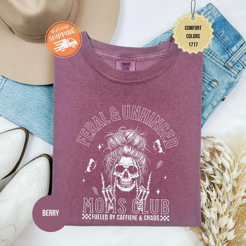 Puede incluir: Sudadera de color bayas con el texto "FERAL & UNHINGED MOMS CLUB" y un gr&aacute;fico de calavera. La camisa tambi&eacute;n tiene el texto "FUELED BY CAFFIENE & CHAOS". La camisa es de la marca Comfort Colors. La imagen incluye un par de jeans, un sombrero y botas.