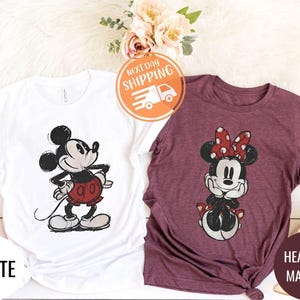 May include: Two t-shirts featuring Mickey and Minnie Mouse. One is white with Mickey in red shorts, the other is heather maroon with Minnie wearing a red polka dot bow. The word "WHITE" is printed on the white shirt.