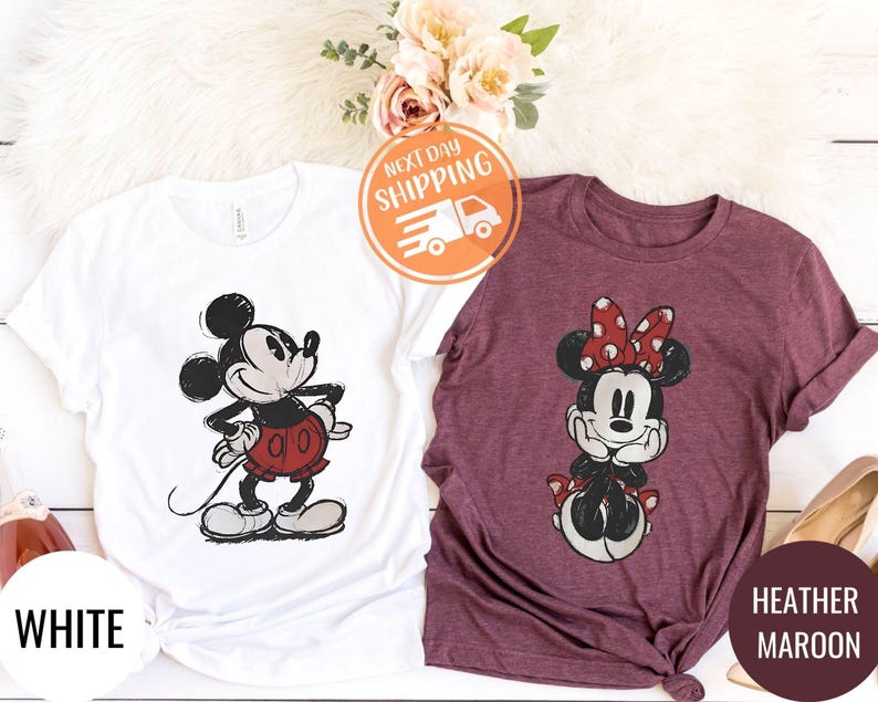 May include: Two t-shirts featuring Mickey and Minnie Mouse. One is white with Mickey in red shorts, the other is heather maroon with Minnie wearing a red polka dot bow. The word "WHITE" is printed on the white shirt.