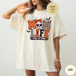 May include: An ivory t-shirt featuring a graphic design with the text MOM LIFE and RAISING TINY HUMANZ. The design includes a skull with a polka dot bow, a spider web, bats, and leopard print patterns in orange, black, and white colors.