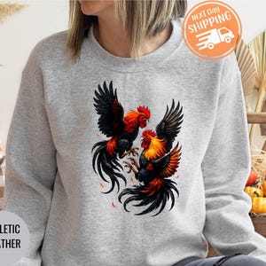 May include: Athletic heather gray sweatshirt featuring a vibrant graphic of two roosters in a fighting pose. The roosters have black and red feathers with orange accents. The sweatshirt has a crew neck and long sleeves.
