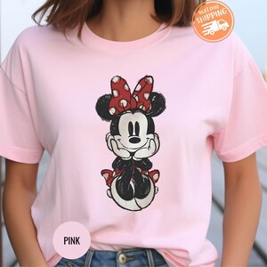 May include: A light pink t-shirt featuring a graphic of Minnie Mouse. Minnie is depicted in black and white with a red polka dot bow. The shirt has short sleeves and a round neckline. The words "Next Day Shipping" are visible.