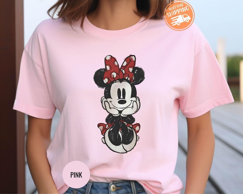 May include: A light pink t-shirt featuring a graphic of Minnie Mouse. Minnie is depicted in black and white with a red polka dot bow. The shirt has short sleeves and a round neckline. The words "Next Day Shipping" are visible.