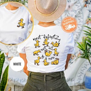 May include: White t-shirt featuring a small yellow duck graphic on the front and a larger design of yellow ducks with the text "most definitely not all in a row" on the back. The shirt is a casual style.