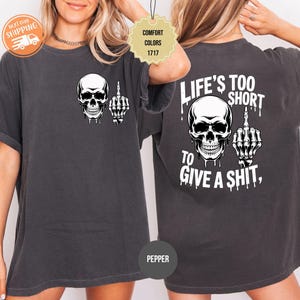 May include: Dark grey t-shirt featuring a white skull graphic with a middle finger gesture. The back of the shirt displays the text "LIFE'S TOO SHORT TO GIVE A SHIT" with a matching skull and hand graphic.