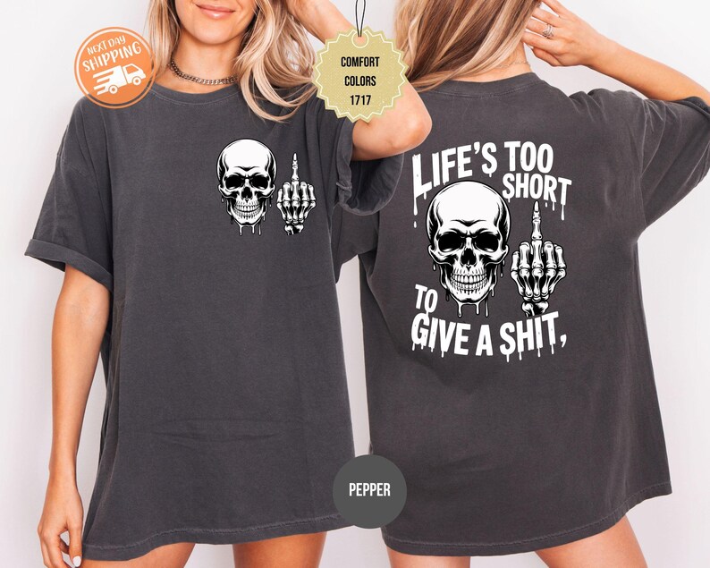 May include: Dark grey t-shirt featuring a white skull graphic with a middle finger gesture. The back of the shirt displays the text "LIFE'S TOO SHORT TO GIVE A SHIT" with a matching skull and hand graphic.