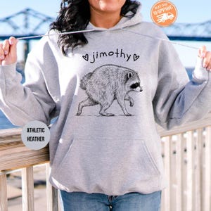 May include: A light gray hooded sweatshirt featuring a black line-art illustration of a raccoon in the center. Above the raccoon, the text reads "jimothy" flanked by two small heart symbols. The garment includes a front kangaroo pocket and drawstrings.