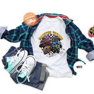 Puede incluir: Camiseta blanca con un gr&aacute;fico colorido de monster trucks y el texto "MONSTER TRUCKS ARE MY JAM". La imagen incluye vaqueros azules, una camisa a cuadros y zapatillas de deporte azul claro.